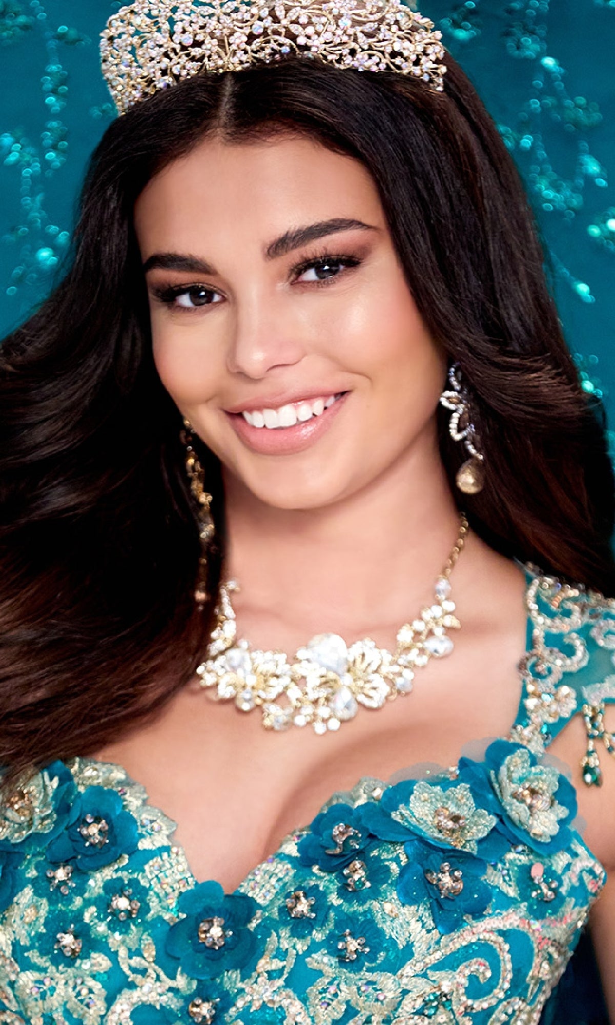 Smiling woman with long dark hair wears the Quinceanera Dress PR30088 By Princesa, a teal lace gown adorned with 3D flowers, jeweled tiara and necklace. Background is turquoise with sparkling details.